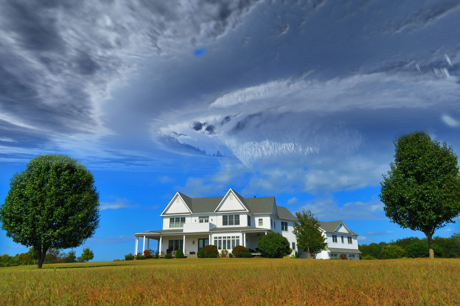 white and gray house between two green tall trees under gray clouds forming swirl during daytime, home