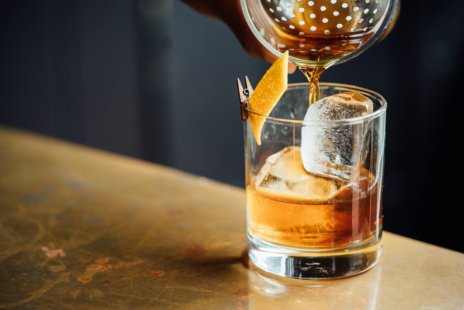Amber liquid pouring from a strainer into a glass with a large ice cube